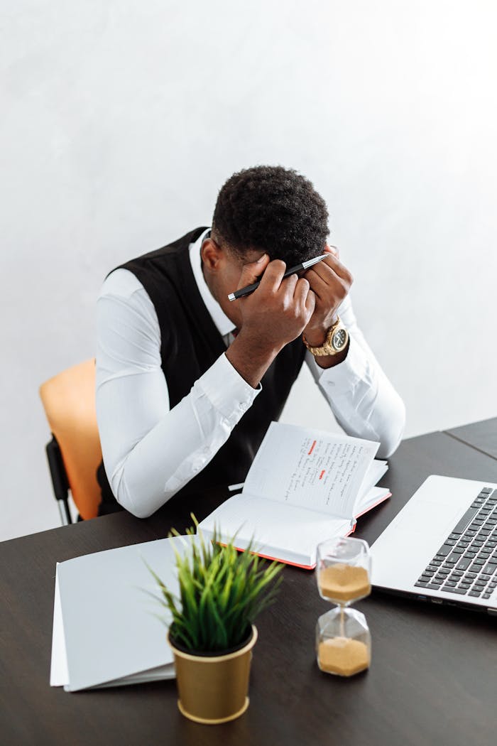 A businessman overwhelmed working indoors with a notebook, laptop, and hourglass.